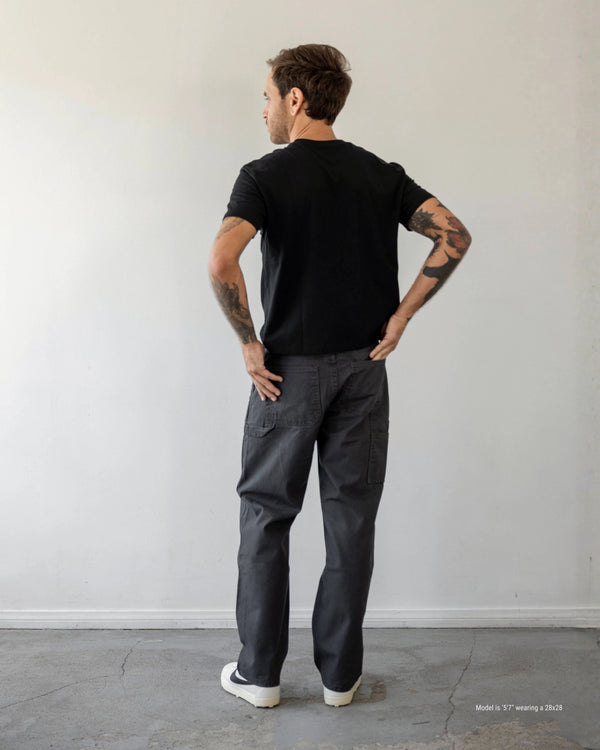 Man wearing a black t-shirt and dark pants standing against a plain white wall.