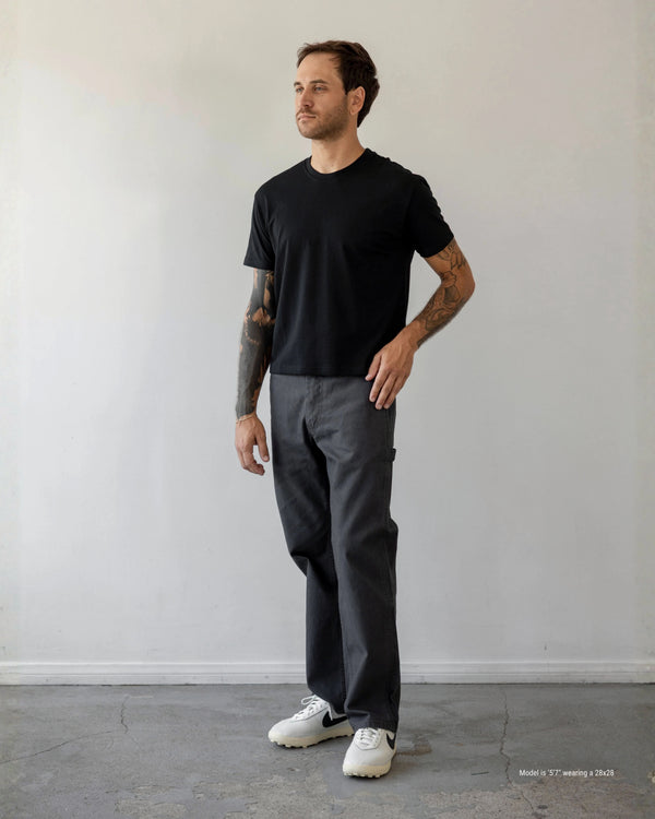 Man wearing a black t-shirt and gray pants standing against a plain white wall.