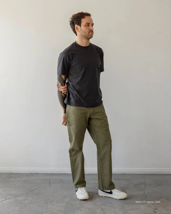 Man wearing a black t-shirt and olive green pants standing against a plain white wall.