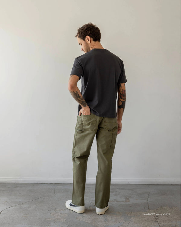 Man wearing a black t-shirt and green cargo pants standing against a plain white wall.