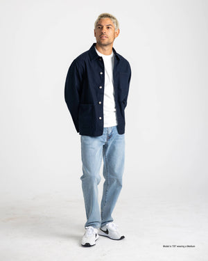 A short man wearing a Stone dark grey Chore Coat over a white T-shirt with black pants and white sneakers, showing the proportional fit against a tan studio background.