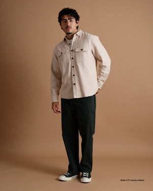 A short male model wearing a tan flannel button-up shirt, dark pants, and black sneakers against a warm brown studio backdrop.