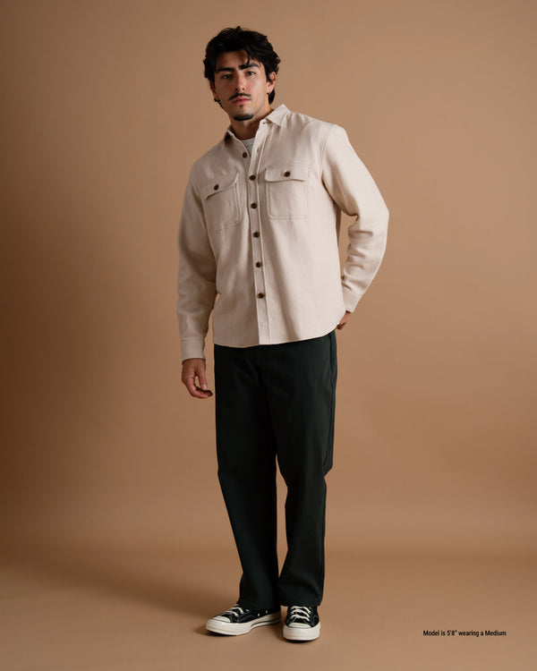 A short male model wearing a tan flannel button-up shirt, dark pants, and black sneakers against a warm brown studio backdrop.