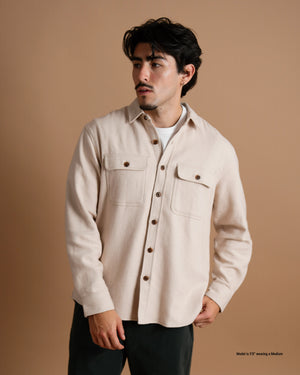 A short male model wearing a tan flannel button-up shirt over a white T-shirt against a warm brown studio backdrop.