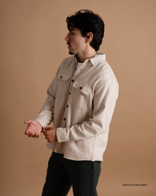 A short male model wearing a tan flannel button-up shirt and dark pants, shown from the side against a warm brown studio backdrop.
