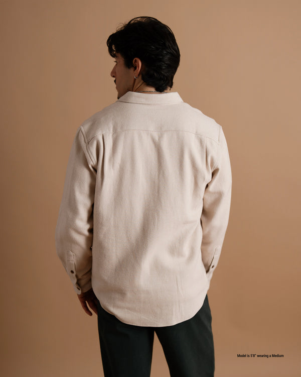 A short male model wearing a tan flannel button-up shirt and dark pants, shown from the back against a warm brown studio backdrop.
