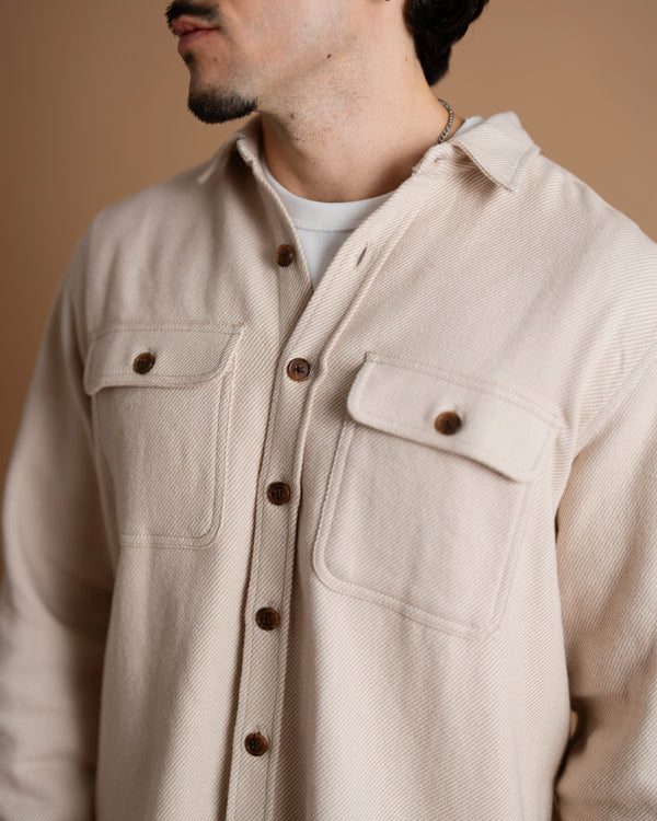 A close-up of a short male model wearing a tan flannel button-up shirt, highlighting the textured fabric, brown buttons, and chest pockets.