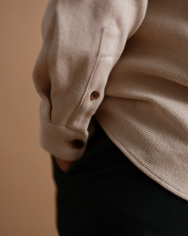 A close-up of a tan flannel shirt worn by a short male model, highlighting the textured fabric, curved hem, and buttoned cuff.