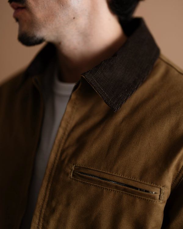 Close-up of a man wearing a brown corduroy jacket with a dark brown corduroy collar and a zippered chest pocket, layered over a white t-shirt.