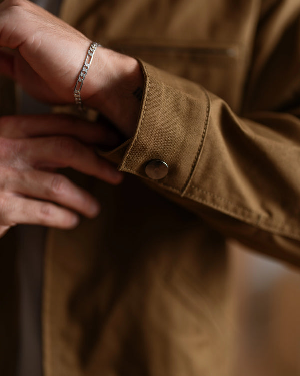 A close-up lifestyle shot of a person's hand adjusting the cuff of a brown canvas utility jacket, wearing a silver chain link bracelet on the wrist.