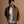 Load image into Gallery viewer, A short man wearing a brown utility-style jacket over a white t-shirt and light blue jeans, standing against a solid warm brown studio background.
