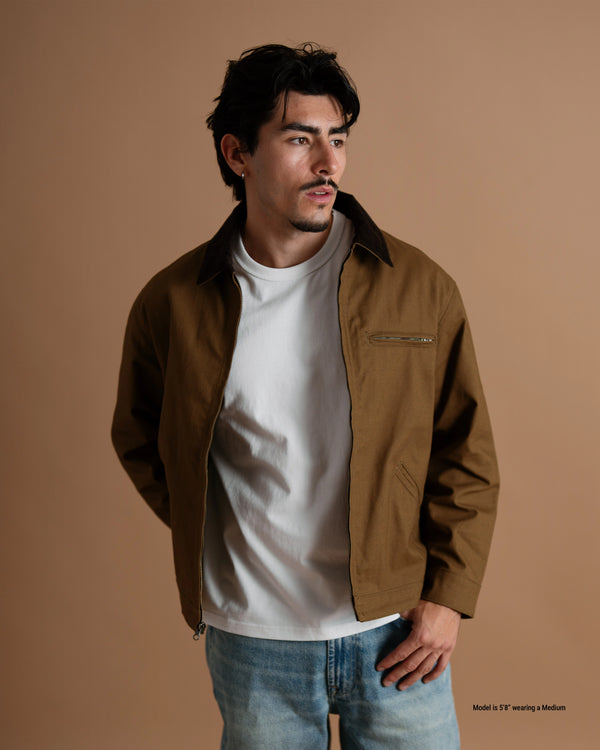 A short man wearing a brown utility-style jacket over a white t-shirt and light blue jeans, standing against a solid warm brown studio background.