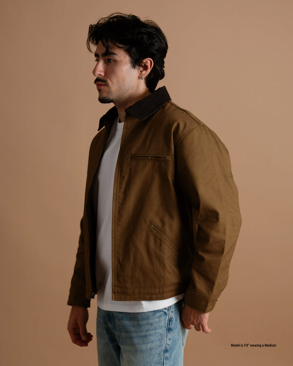 A 5'8" man wearing a brown canvas utility jacket with a dark collar, layered over a white t-shirt and blue jeans, standing against a solid warm brown studio background.