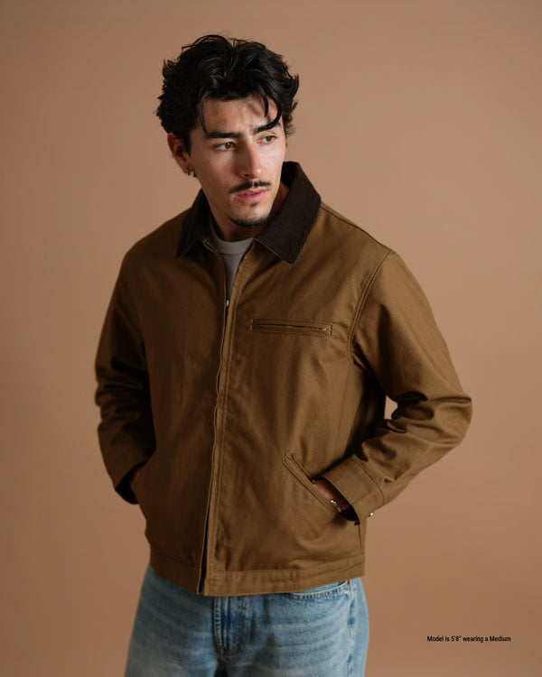 A man wearing a brown utility jacket with a dark brown corduroy collar, paired with light blue jeans, standing against a solid warm brown studio background.