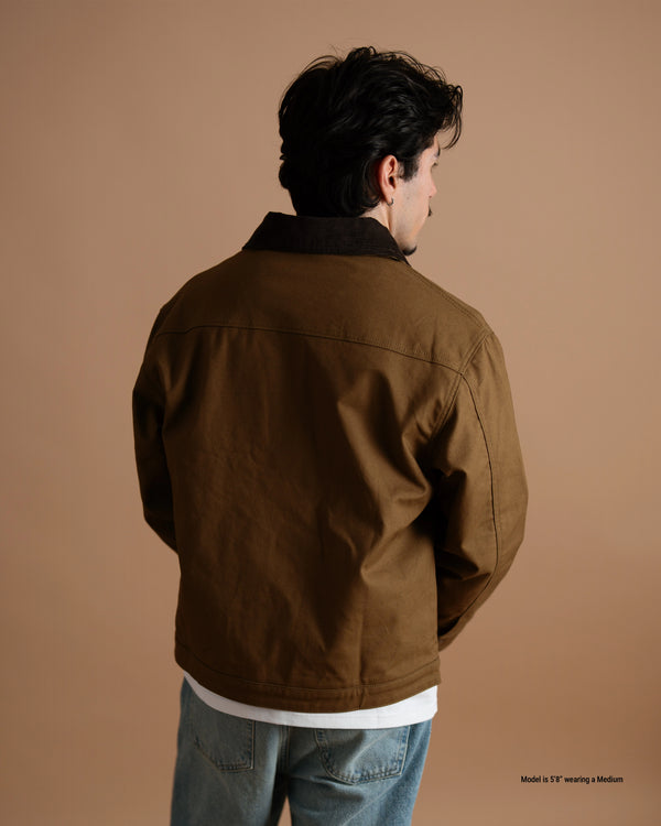 A man shown from the back wearing a brown jacket with a black collar, blue jeans, and a white undershirt, set against a solid warm brown studio background.