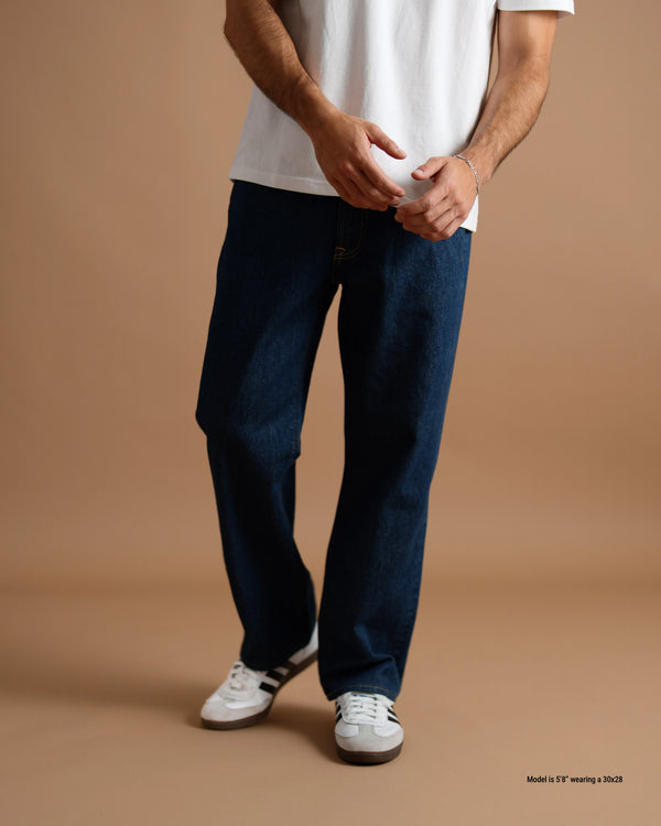 A model wearing dark blue straight-leg denim jeans, a white t-shirt, and white sneakers, standing against a solid tan studio backdrop.