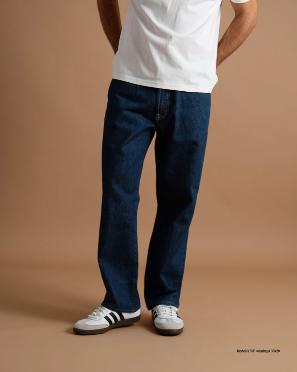 A person wearing dark blue straight-leg jeans, a white t-shirt, and white sneakers with black stripes, standing against a solid tan studio background.