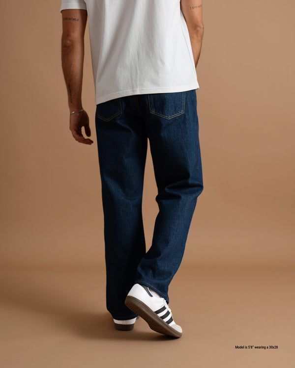 A person shown from the back wearing dark blue straight-leg jeans, a white t-shirt, and white sneakers with black stripes, standing against a solid tan studio background.