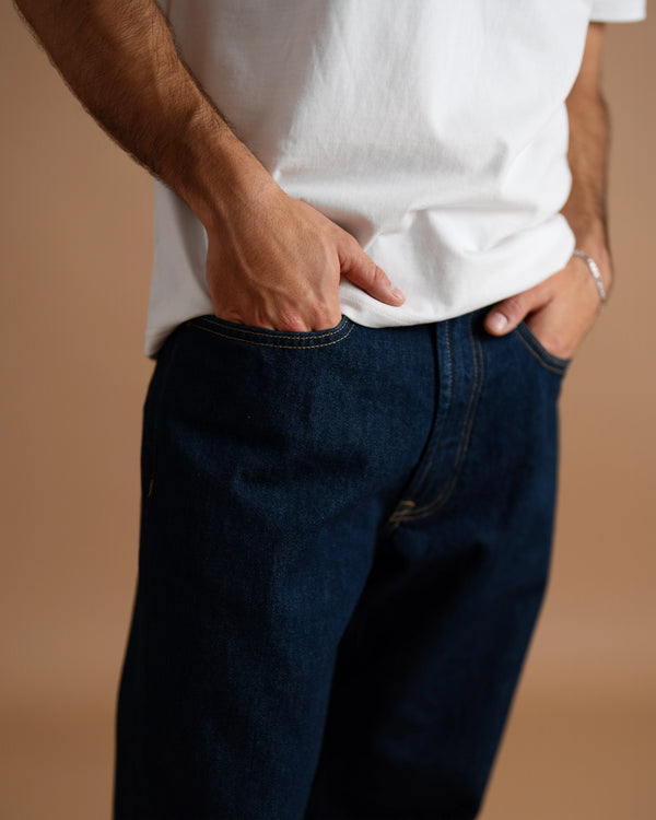 A close-up of a person wearing dark indigo blue jeans and a white t-shirt, with hands in the pockets, set against a solid warm brown background.