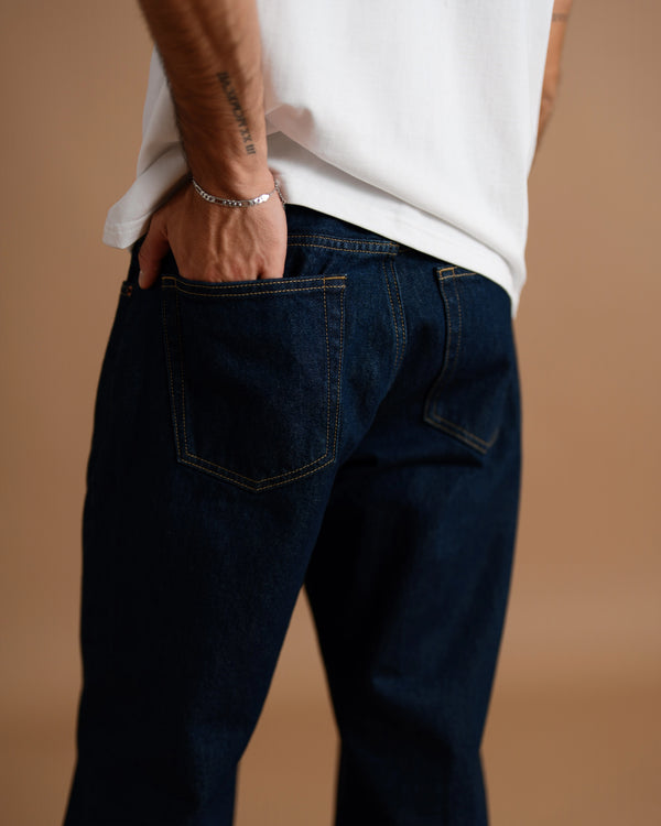 A close-up back view of a person wearing dark indigo denim jeans with a white t-shirt, standing against a solid warm beige background.