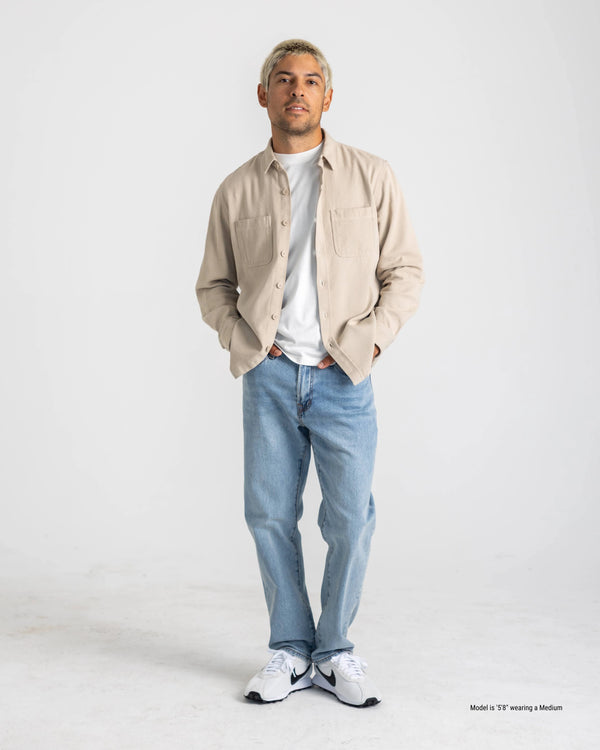 Short Man wearing a beige shirt, white t-shirt, light blue jeans, and white sneakers on a white background