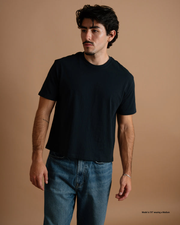 A man wearing a black short-sleeve crewneck t-shirt and blue jeans, standing against a solid warm brown studio background.