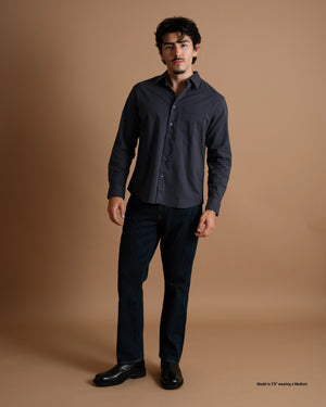 A short man wearing a dark navy Obsidian Washed Lightweight Long-Sleeve Shirt with dark jeans and black shoes, shown from the front against a warm brown studio background.
