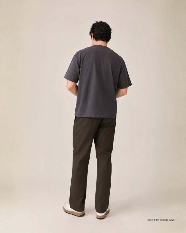 A man viewed from behind wearing a dark charcoal gray short-sleeve t-shirt and dark olive brown pants, standing against a solid light beige studio background.