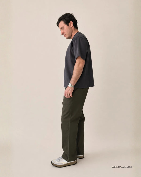 A man stands in profile wearing a dark charcoal gray short-sleeve T-shirt, olive green trousers, and white sneakers against a plain beige studio background.