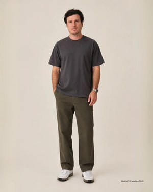A man stands against a plain warm beige studio background wearing a dark charcoal gray short-sleeve crewneck t-shirt, olive green pants, and white sneakers. Small text in the bottom right corner reads 'Model wearing a 32x28'.