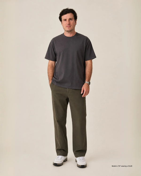 A man stands against a plain warm beige studio background wearing a dark charcoal gray short-sleeve crewneck t-shirt, olive green pants, and white sneakers. Small text in the bottom right corner reads 'Model wearing a 32x28'.