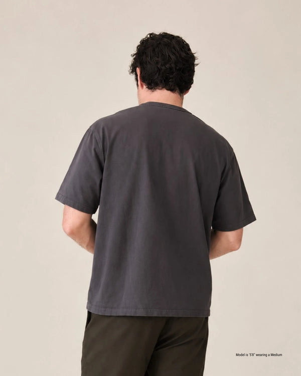 A man viewed from behind wearing a dark charcoal gray short-sleeve t-shirt and dark brown pants, standing against a plain beige studio background.