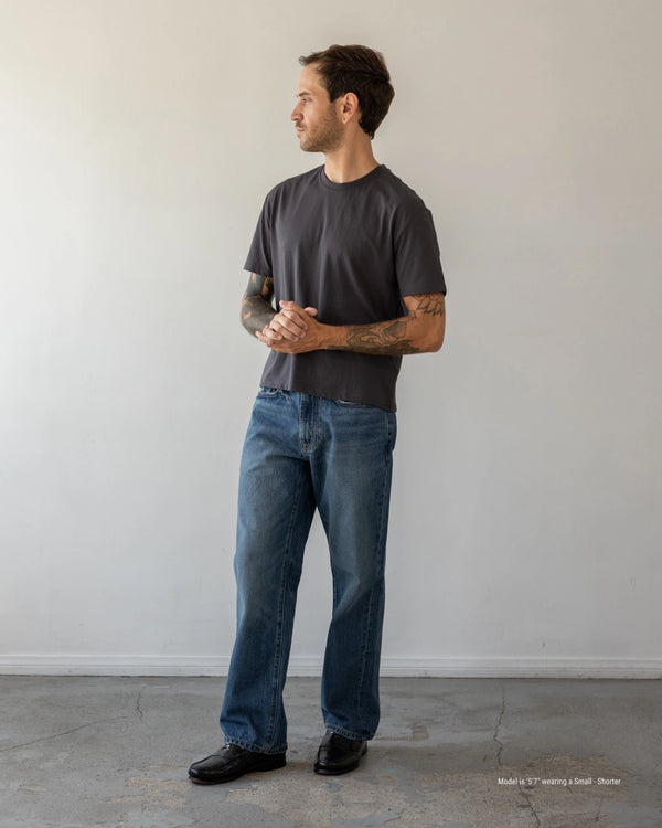 Man wearing a dark gray t-shirt and blue jeans standing against a plain white wall.