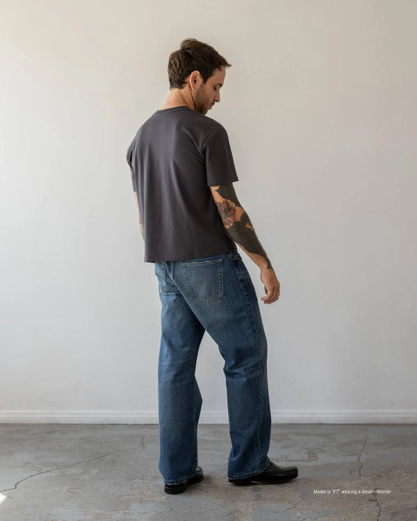 Man wearing a dark t-shirt and blue jeans standing against a plain white wall.