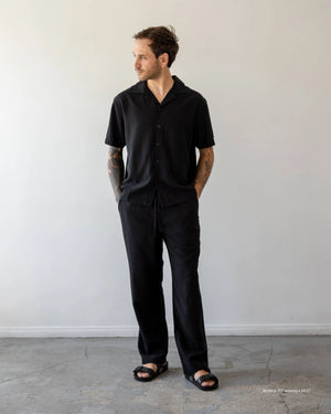 Man wearing a black outfit standing against a plain white wall.