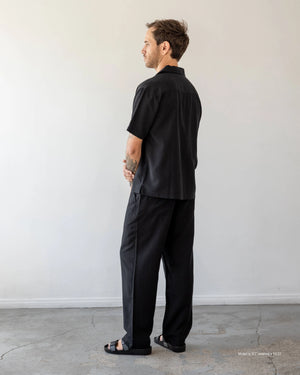 A short male model shown from the back wearing black Tumbled Linen Loose Trousers with a black short-sleeve shirt and sandals against a white studio wall.
