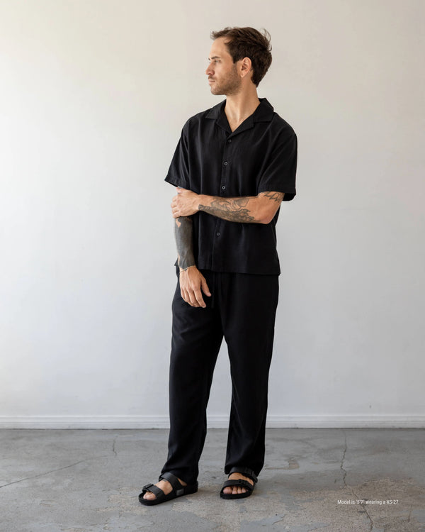 Man wearing a black outfit standing against a plain white wall.