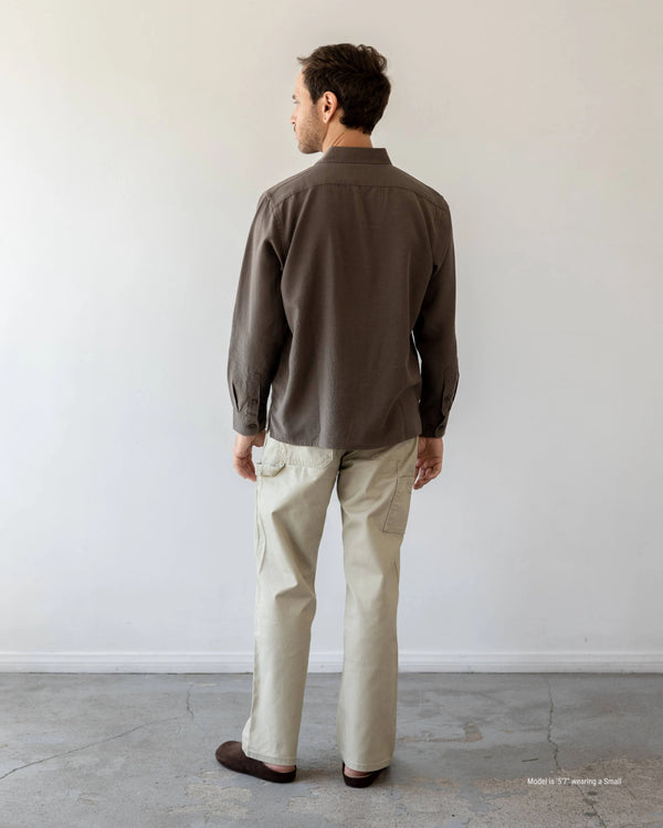 Man wearing a brown shirt and beige pants standing against a plain white wall.