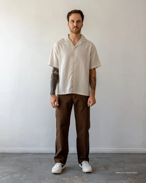 Shorter Man wearing a brown shirt and beige pants standing against a white wall.