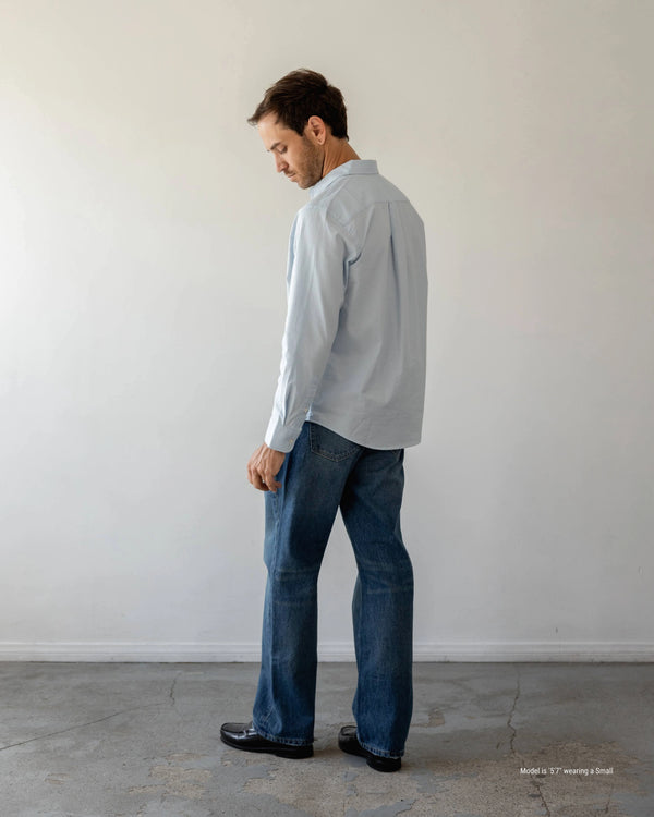 Man wearing a light blue shirt and dark jeans standing against a plain white wall.