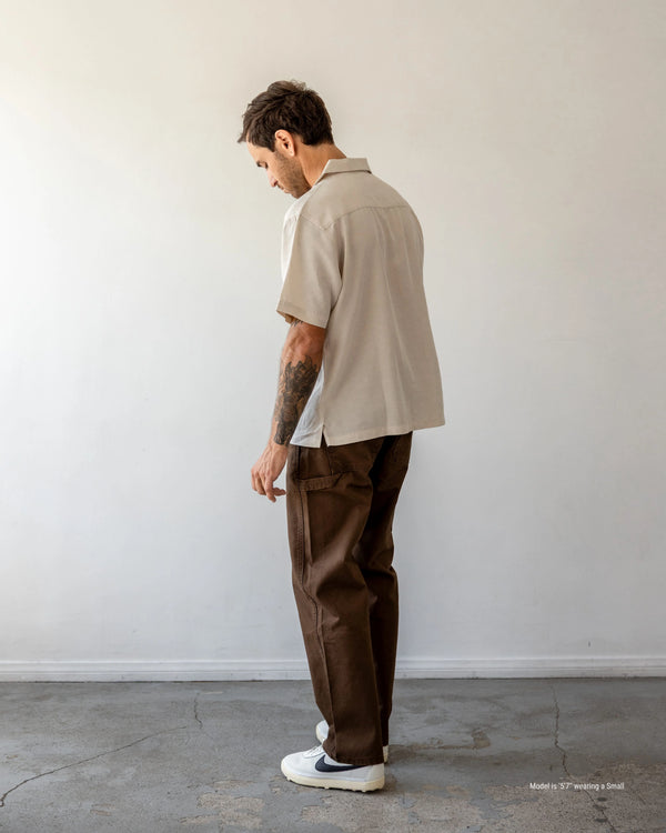 Man wearing a beige shirt and brown pants standing against a white wall.