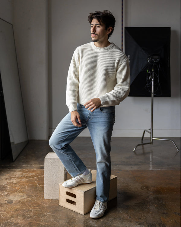 Short Man in a white sweater and blue jeans standing on a wooden box in a studio setting.