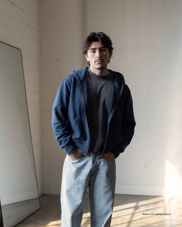 Man wearing a blue hoodie and gray pants standing in a minimalistic room.