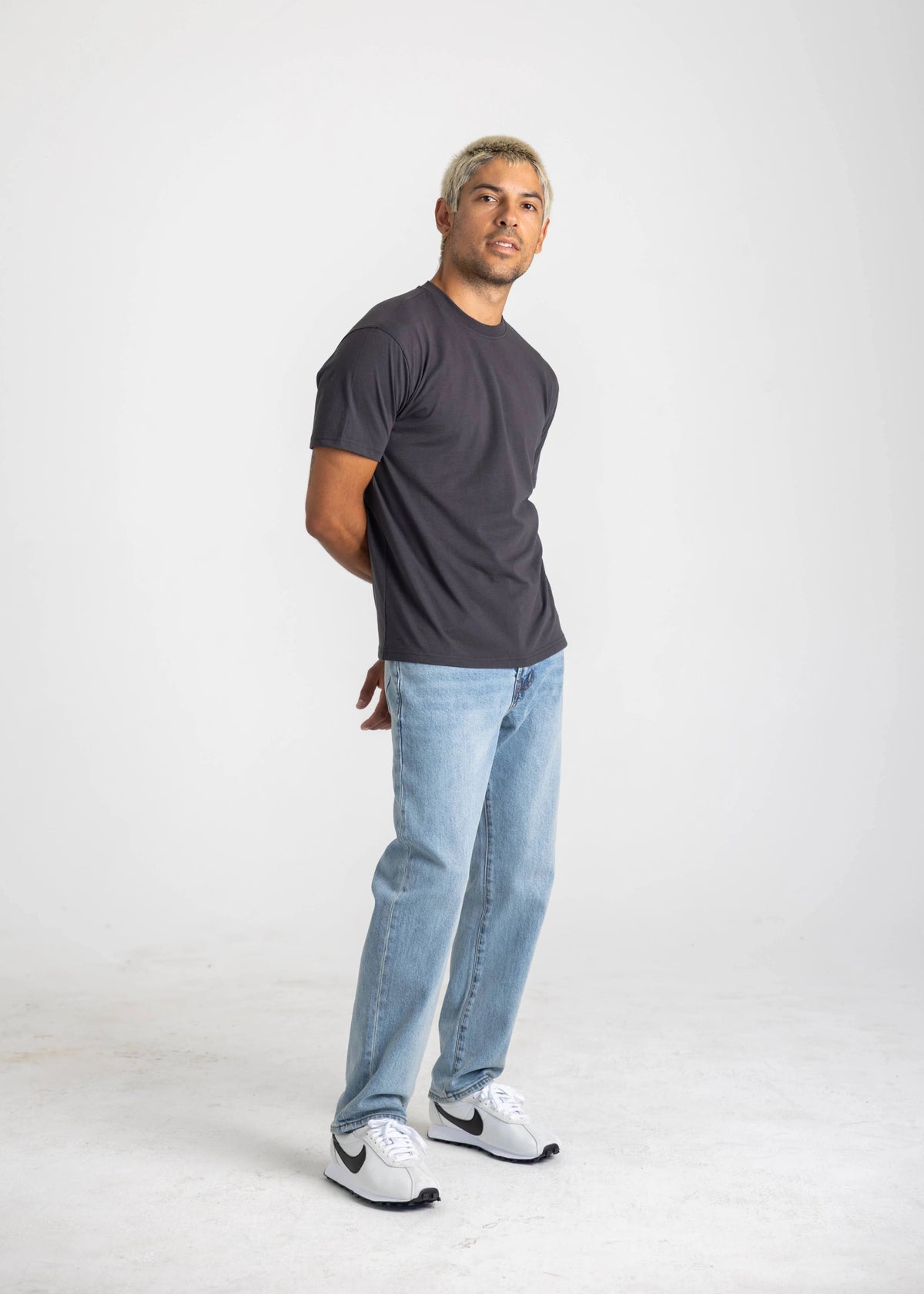 Man wearing a dark t-shirt, light jeans, and white sneakers on a white background