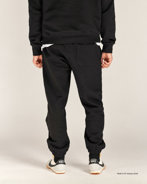 Back view of black jogger sweatpants on a shorter man, showing the back pocket, tapered leg and elastic cuffs
