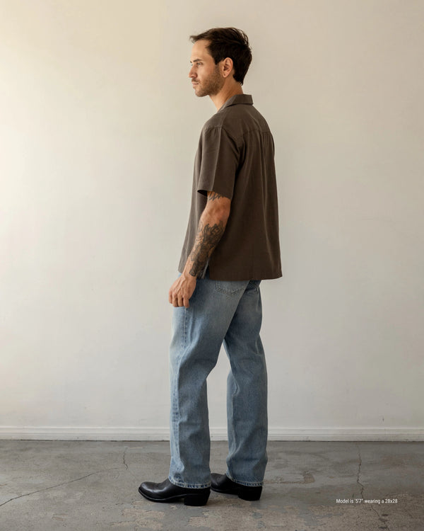 Shorter Man wearing a brown shirt and blue jeans standing against a plain white wall.