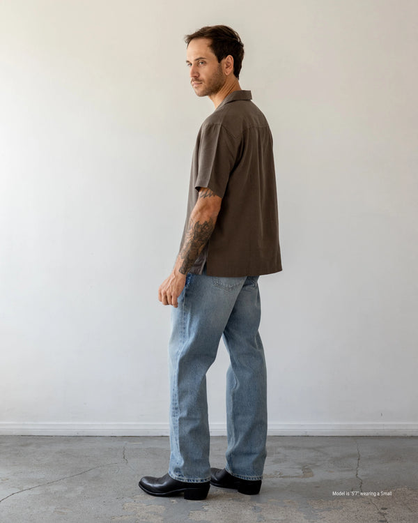 Man wearing a brown shirt and blue jeans standing against a white wall.