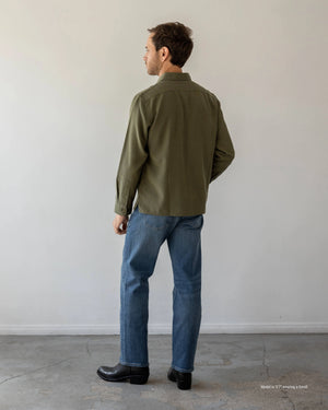 A short male model shown from the back wearing a long-sleeve olive green Tumbled Linen Shirt with blue jeans against a plain white studio background.
