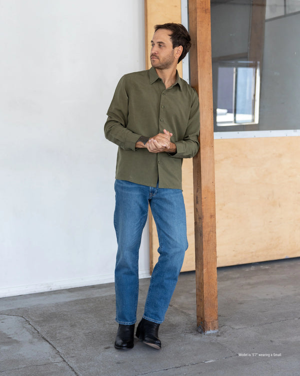 Man wearing a green shirt and blue jeans standing against a white wall.