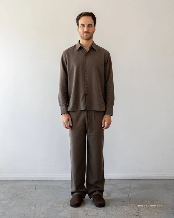 Man wearing a brown shirt and pants standing against a white wall.